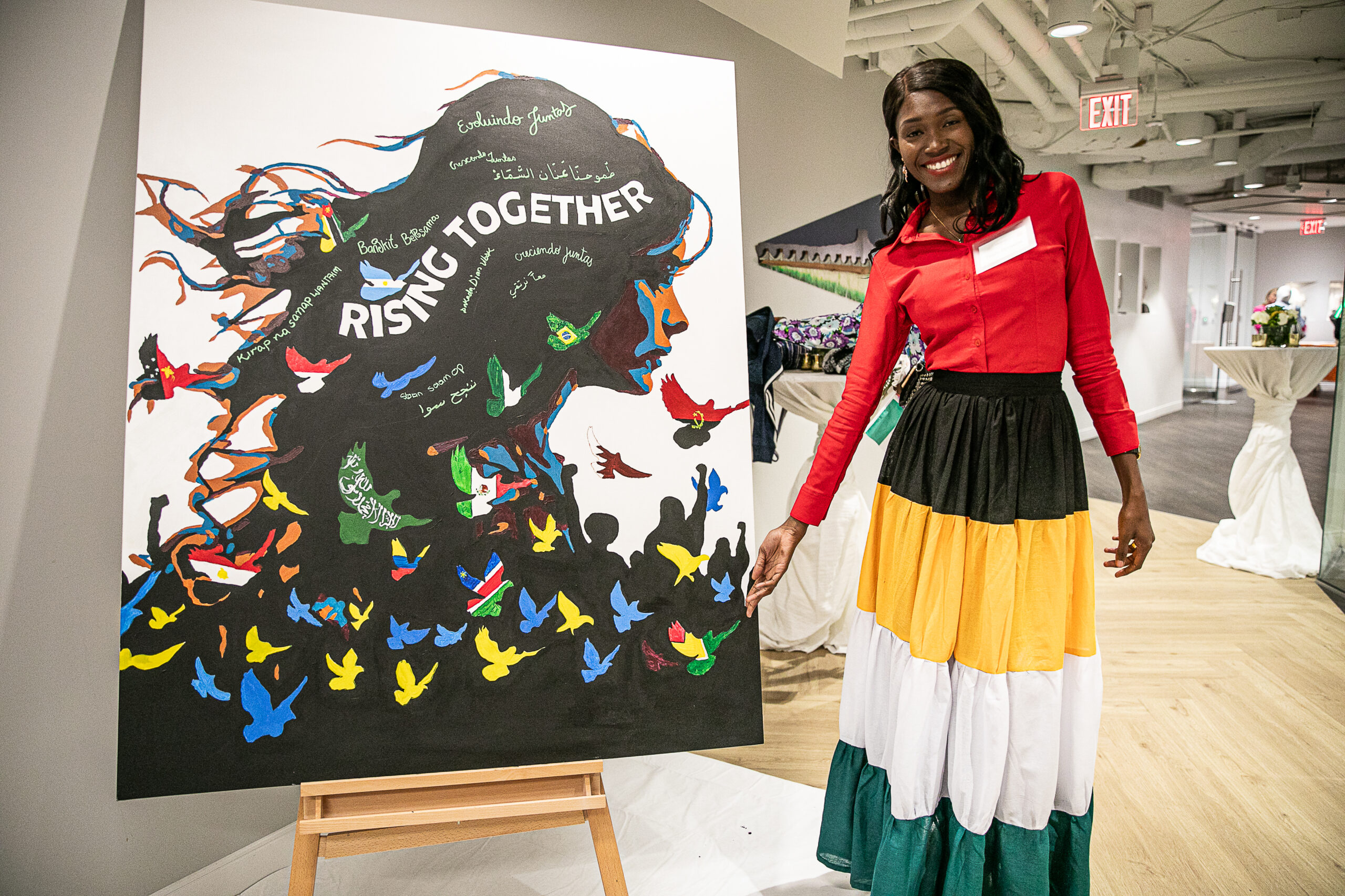Tashi Browne posing with artwork created by Global Women In Management alumni, featuring the message "Rising Together" and birds painted to represent the flags of alumni home countries.